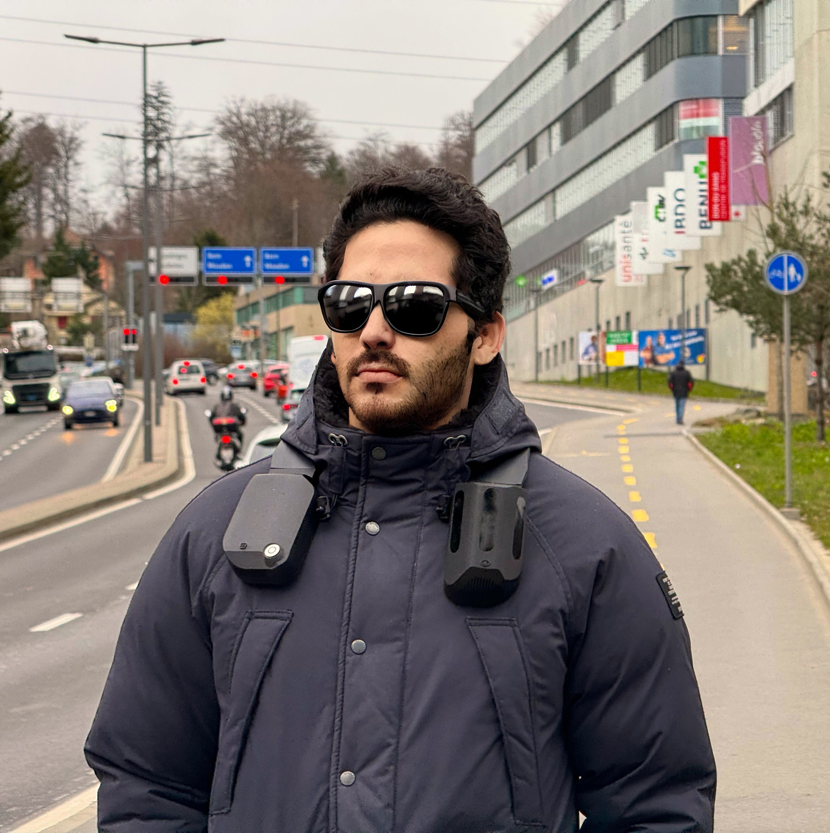 A man wearing a modern dark down jacket with a gadget attached to his shoulders, wearing dark sunglasses, and standing on a city sidewalk with a road and city traffic in the background. He uses this device in his daily life. The text next to his review reads: 'I honestly don't think I've ever bought something with so much potential to change lives. - Stuart Beveridge from Seescape.